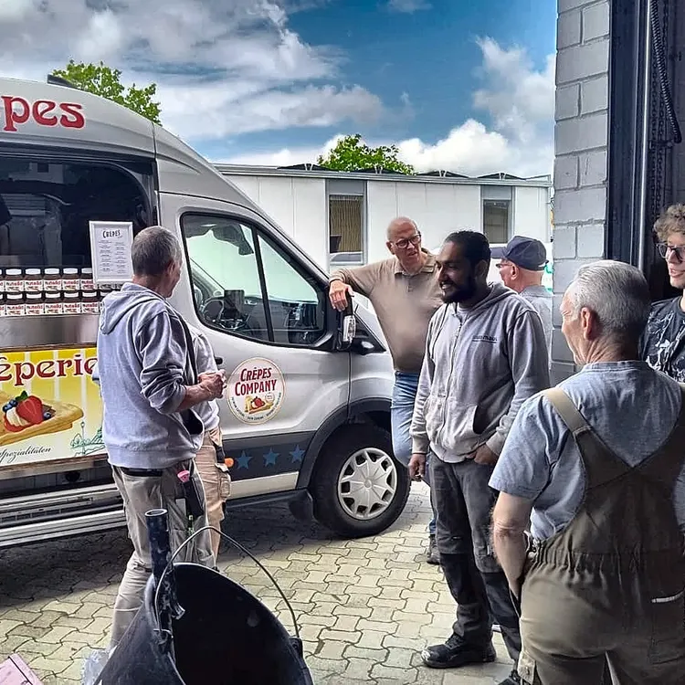 Mitarbeitende stehen im Hof vor einem geöffneten Crêpes-Wagen und warten auf ihre Bestellung.