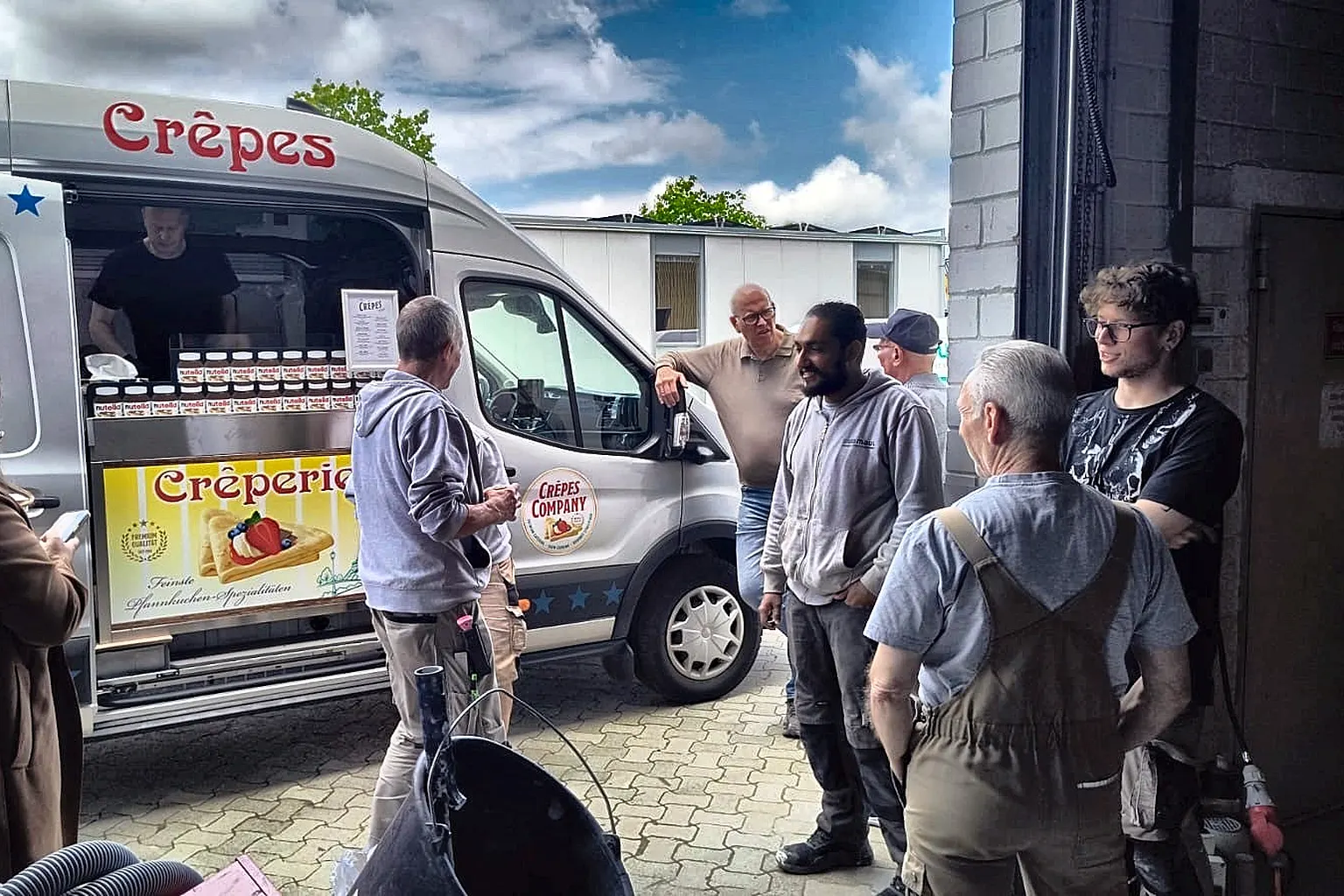 Mitarbeitende stehen im Hof vor einem geöffneten Crêpes-Wagen und warten auf ihre Bestellung.